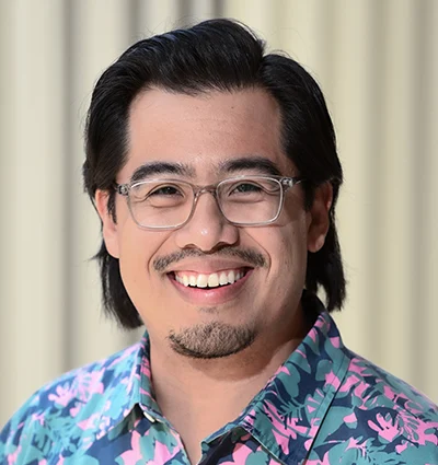 A person with long dark hair, glasses, and facial hair is smiling. They are wearing a blue shirt with a colorful pattern. The background shows vertical blinds in soft focus.