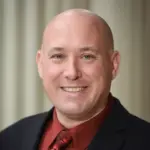 A person with a shaved head is smiling, wearing a dark suit, a red shirt, and a patterned tie. The background is softly blurred, likely indoors.