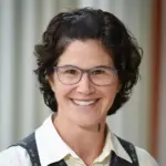 Person with short, curly dark hair, wearing glasses, a white shirt, and a dark vest, smiling against a blurred background.