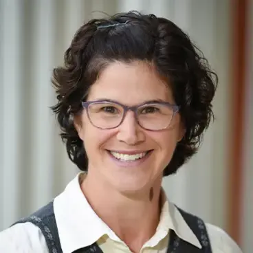 Person with short, curly dark hair, wearing glasses, a white shirt, and a dark vest, smiling against a blurred background.