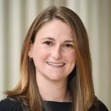 A woman with long, straight brown hair wearing a black top smiles at the camera, standing in front of a light-colored, blurred background.
