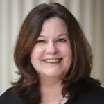 A person with shoulder-length dark hair smiles at the camera. They are wearing a black top and a silver necklace. The background is softly blurred with vertical lines.