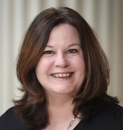 A person with shoulder-length dark hair smiles at the camera. They are wearing a black top and a silver necklace. The background is softly blurred with vertical lines.