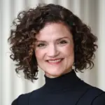 Smiling person with curly hair wearing a black turtleneck, standing in front of a neutral background.