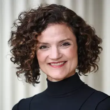 Smiling person with curly hair wearing a black turtleneck, standing in front of a neutral background.