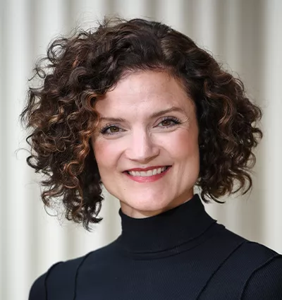 Smiling person with curly hair wearing a black turtleneck, standing in front of a neutral background.