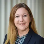 A woman with long, light brown hair smiles at the camera. She is wearing a dark blazer and a patterned blouse, standing against a blurred, light-colored background.
