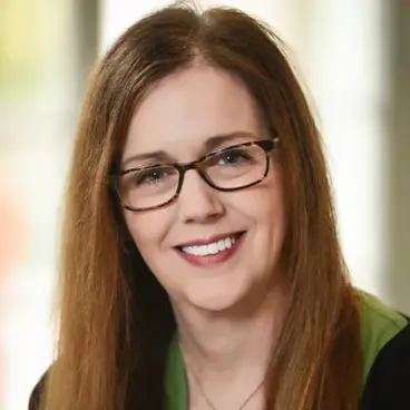 A woman with long brown hair and glasses smiles at the camera. She is wearing a dark top with a green collar. The background is softly blurred, creating a warm, natural atmosphere.