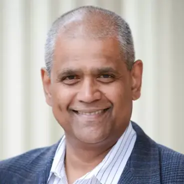 Smiling person with short hair and a gray suit jacket over a striped shirt, standing in front of a neutral background.