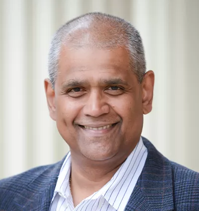 Smiling person with short hair and a gray suit jacket over a striped shirt, standing in front of a neutral background.