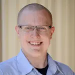 A smiling person with glasses and a shaved head, wearing a light blue collared shirt, stands in front of a blurred beige background.