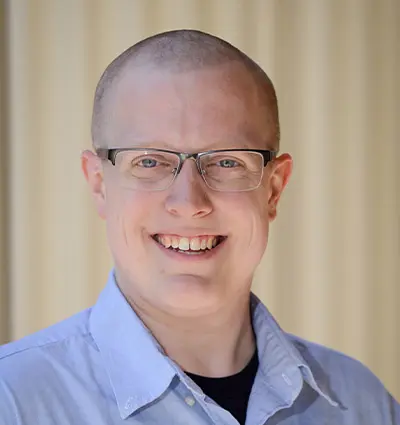 A smiling person with glasses and a shaved head, wearing a light blue collared shirt, stands in front of a blurred beige background.