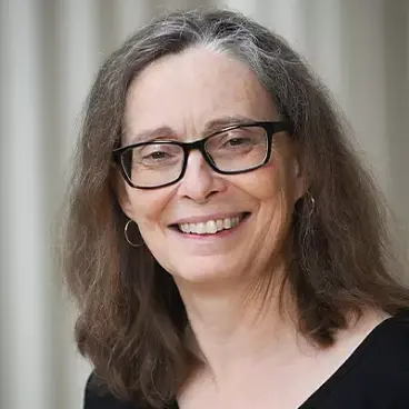 A smiling woman with long brown hair and glasses wearing a black top and hoop earrings.