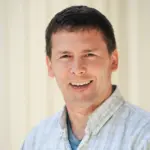 Smiling person with short dark hair, wearing a light plaid shirt over a blue t-shirt, standing against a light-colored background.