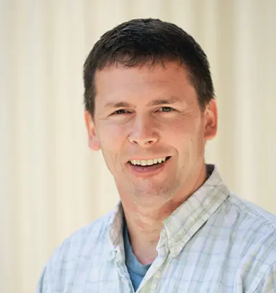 Smiling person with short dark hair, wearing a light plaid shirt over a blue t-shirt, standing against a light-colored background.