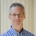 A person with short gray hair and glasses is wearing a blue and white checkered shirt. They are standing in front of a cream-colored background and are looking at the camera with a neutral expression.