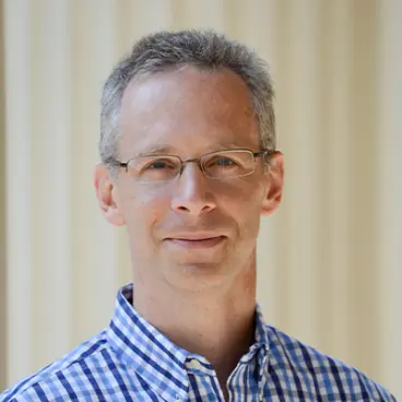 A person with short gray hair and glasses is wearing a blue and white checkered shirt. They are standing in front of a cream-colored background and are looking at the camera with a neutral expression.