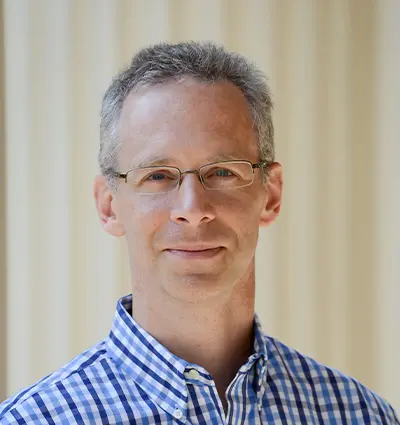 A person with short gray hair and glasses is wearing a blue and white checkered shirt. They are standing in front of a cream-colored background and are looking at the camera with a neutral expression.