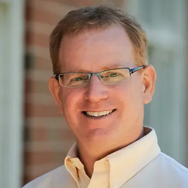 A man with short hair and glasses is smiling in an outdoor setting. He is wearing a light-colored button-up shirt. The background features a brick wall and a window.