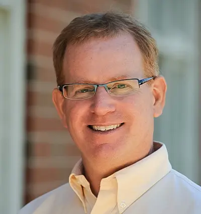 A man with short hair and glasses is smiling in an outdoor setting. He is wearing a light-colored button-up shirt. The background features a brick wall and a window.