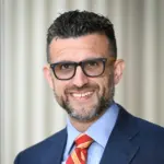 Smiling person with short, dark curly hair and a beard, wearing glasses, a blue suit, light blue shirt, and a red striped tie, standing in front of a neutral, blurred background.
