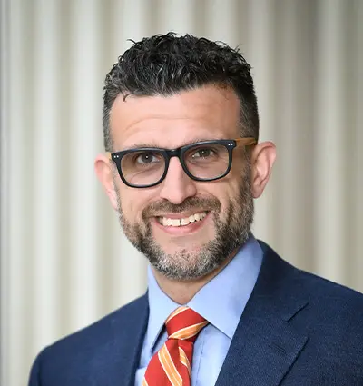 Smiling person with short, dark curly hair and a beard, wearing glasses, a blue suit, light blue shirt, and a red striped tie, standing in front of a neutral, blurred background.