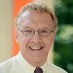A man with curly light hair and glasses smiles at the camera. He is wearing a light-colored shirt and a tie. The background is softly blurred, showing hints of greenery and an orange vertical stripe.