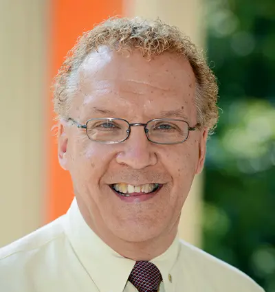 A man with curly light hair and glasses smiles at the camera. He is wearing a light-colored shirt and a tie. The background is softly blurred, showing hints of greenery and an orange vertical stripe.
