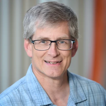 A man with short gray hair and glasses smiling at the camera. He is wearing a plaid shirt and standing in front of a light-colored curtain.