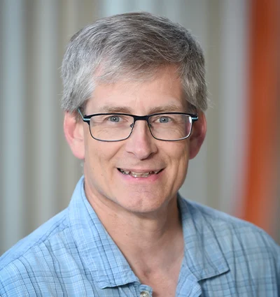 A man with short gray hair and glasses smiling at the camera. He is wearing a plaid shirt and standing in front of a light-colored curtain.