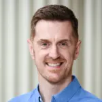 Smiling person with short brown hair, wearing a blue button-up shirt, stands in front of a blurred neutral background.