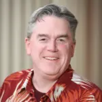 A man smiling while wearing a red and white patterned shirt. He has short gray hair and is standing against a plain, blurred background.