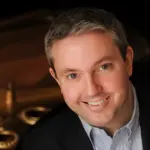 Smiling man with short gray hair, wearing a dark jacket and light blue shirt, sits indoors with a blurred background.