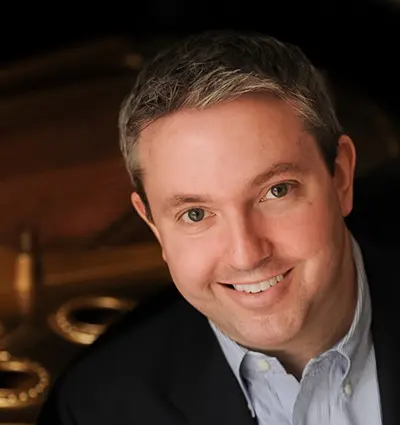 Smiling man with short gray hair, wearing a dark jacket and light blue shirt, sits indoors with a blurred background.