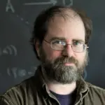 A bearded man with glasses stands in front of a chalkboard covered with diagrams and mathematical symbols. He has a contemplative expression and wears a brown shirt over a black t-shirt.
