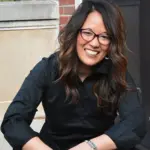 A person with long dark hair and glasses is smiling while wearing a black shirt. They are seated outdoors in front of a brick and concrete wall.