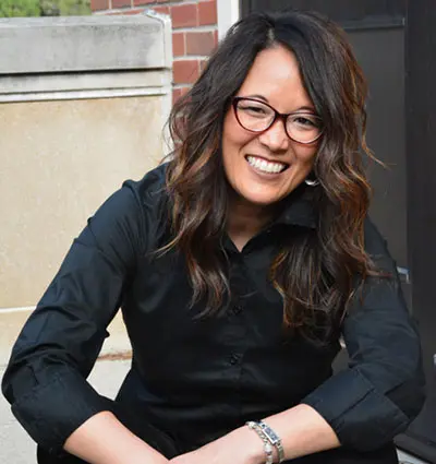 A person with long dark hair and glasses is smiling while wearing a black shirt. They are seated outdoors in front of a brick and concrete wall.
