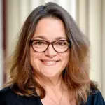 A person with long brown hair, wearing glasses and a black top, smiles against a light-colored curtain backdrop.