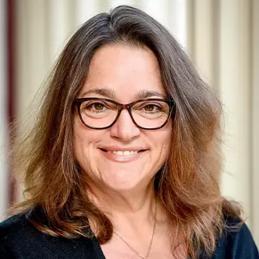 A person with long brown hair, wearing glasses and a black top, smiles against a light-colored curtain backdrop.