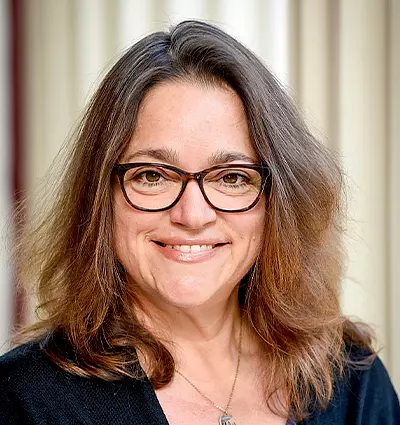 A person with long brown hair, wearing glasses and a black top, smiles against a light-colored curtain backdrop.