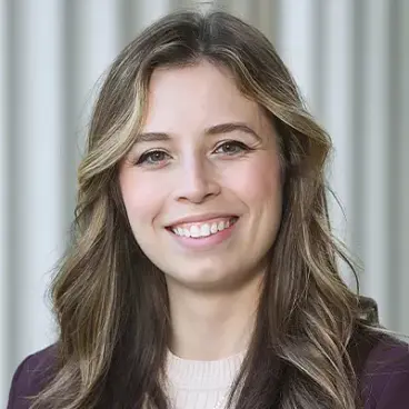 A woman with long wavy hair smiles at the camera. She is wearing a dark blazer over a light top. The background features vertical gray stripes.
