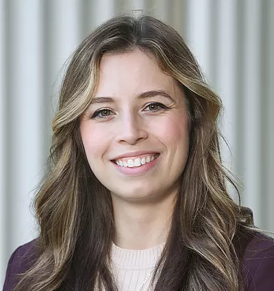 A woman with long wavy hair smiles at the camera. She is wearing a dark blazer over a light top. The background features vertical gray stripes.