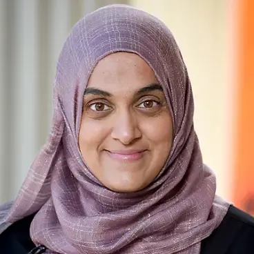 A person wearing a purple hijab smiles at the camera. The background is softly blurred, featuring warm colors.
