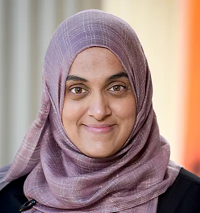 A person wearing a purple hijab smiles at the camera. The background is softly blurred, featuring warm colors.