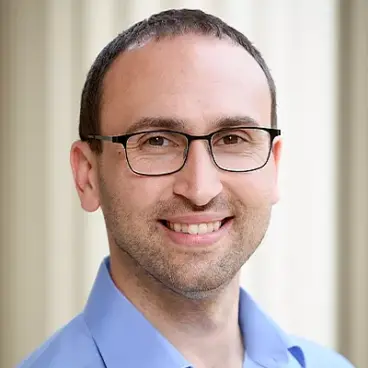 A person with short dark hair and glasses, smiling while wearing a light blue shirt. The background is softly blurred, giving a professional and friendly appearance.