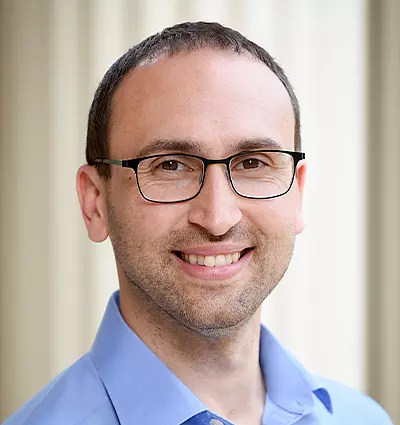 A person with short dark hair and glasses, smiling while wearing a light blue shirt. The background is softly blurred, giving a professional and friendly appearance.