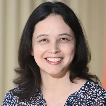 A person with long black hair is smiling, wearing a black top with floral patterns. The background is softly blurred in neutral tones.