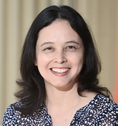 A person with long black hair is smiling, wearing a black top with floral patterns. The background is softly blurred in neutral tones.