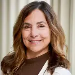 A woman with long, wavy brown hair smiles while wearing a white sweater over a brown turtleneck. The background features vertical cream-colored blinds.