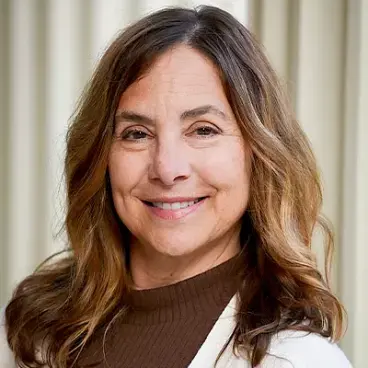 A woman with long, wavy brown hair smiles while wearing a white sweater over a brown turtleneck. The background features vertical cream-colored blinds.